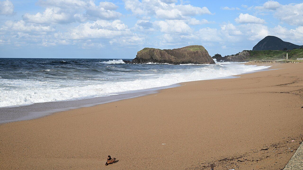 A wide sandy beach on the Sea of Japan coast at Taiza in Kyotango, with rolling surf and the Tateiwa rock under a blue sky